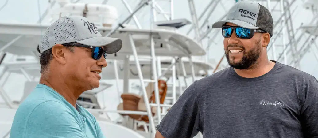 Two men wearing sunglasses and caps, standing in front of a boat.