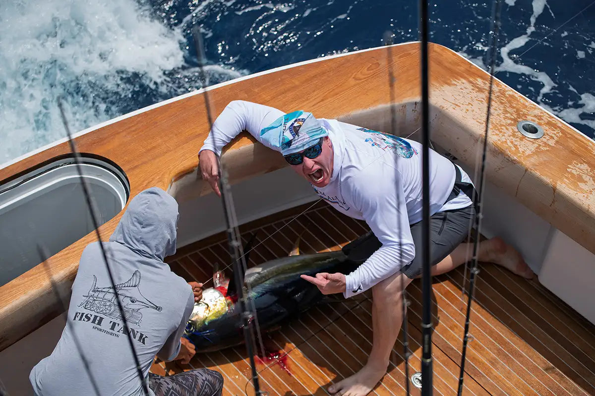 Two men on a boat with fishing rods, one holding a large fish wearing hook optics